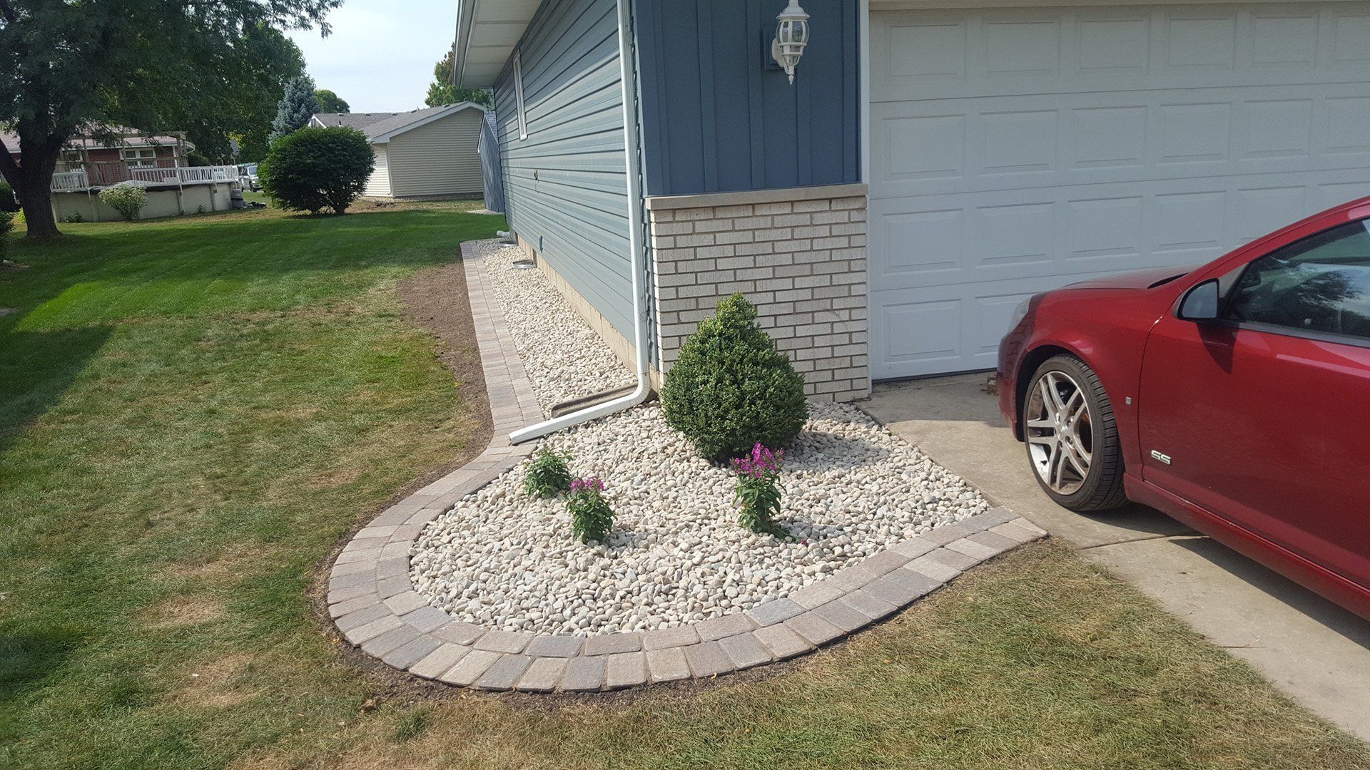 Red car parked beside a house with blue siding, a stone facade, and a landscaped bed with rocks and plants.