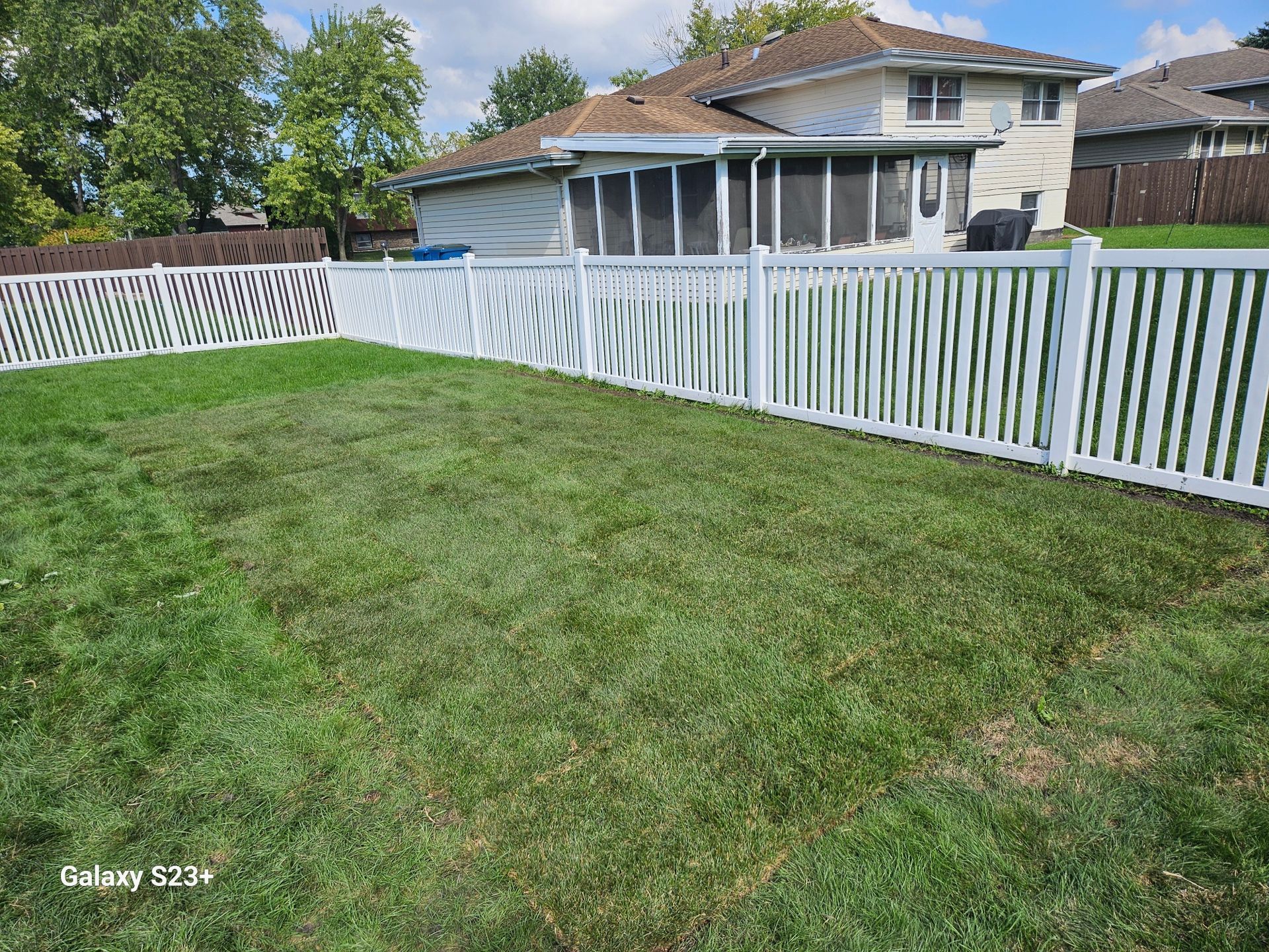 White picket fence surrounds a green backyard, adjacent to a house under a blue sky.