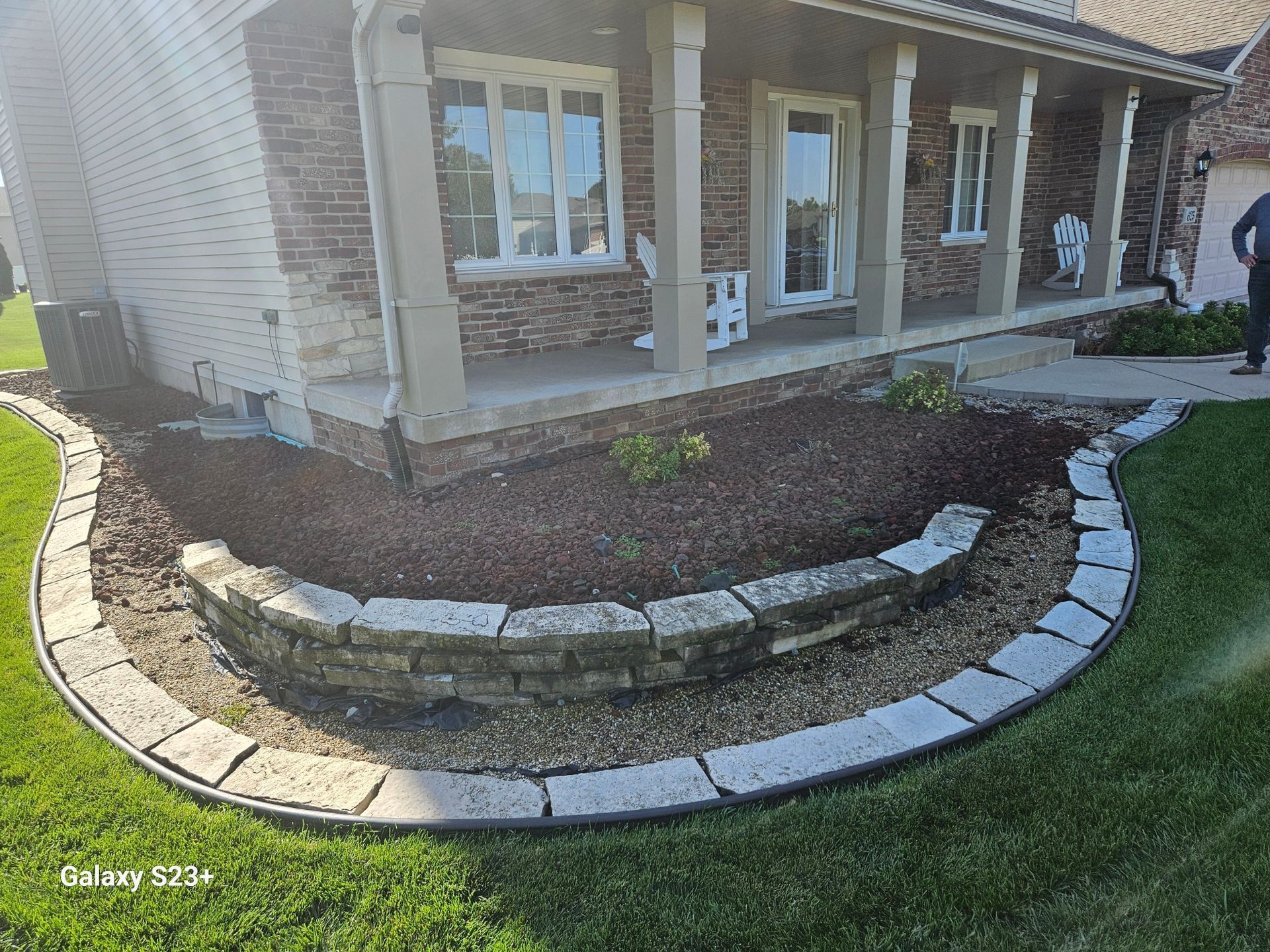 Stone-edged flower bed in front of a brick house with a porch and columns, filled with mulch and plants.