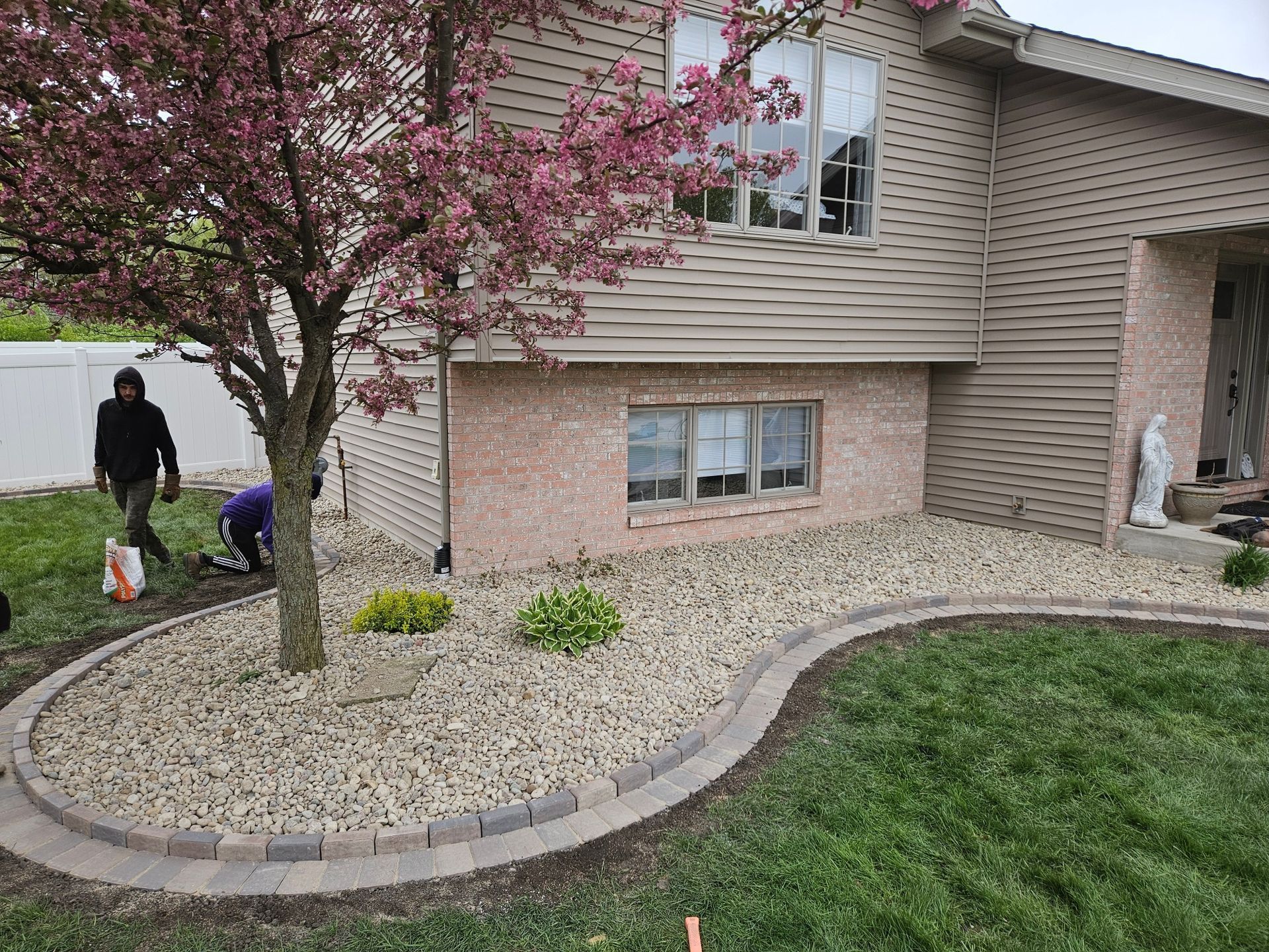 Landscaped yard with tree, gravel bed, and brick border; people working.