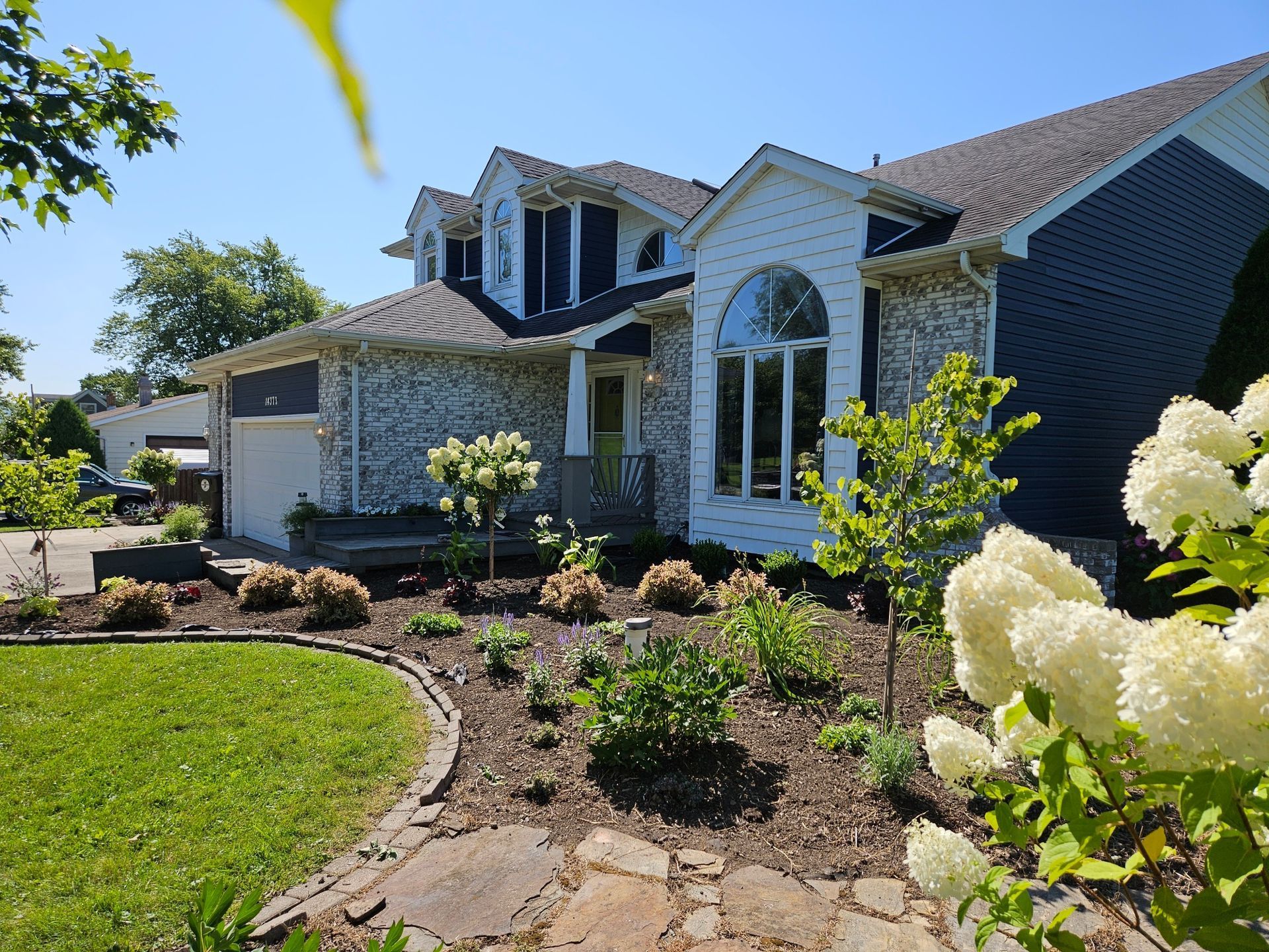 House with light stone and blue siding, arched windows, and a landscaped front yard on a sunny day.