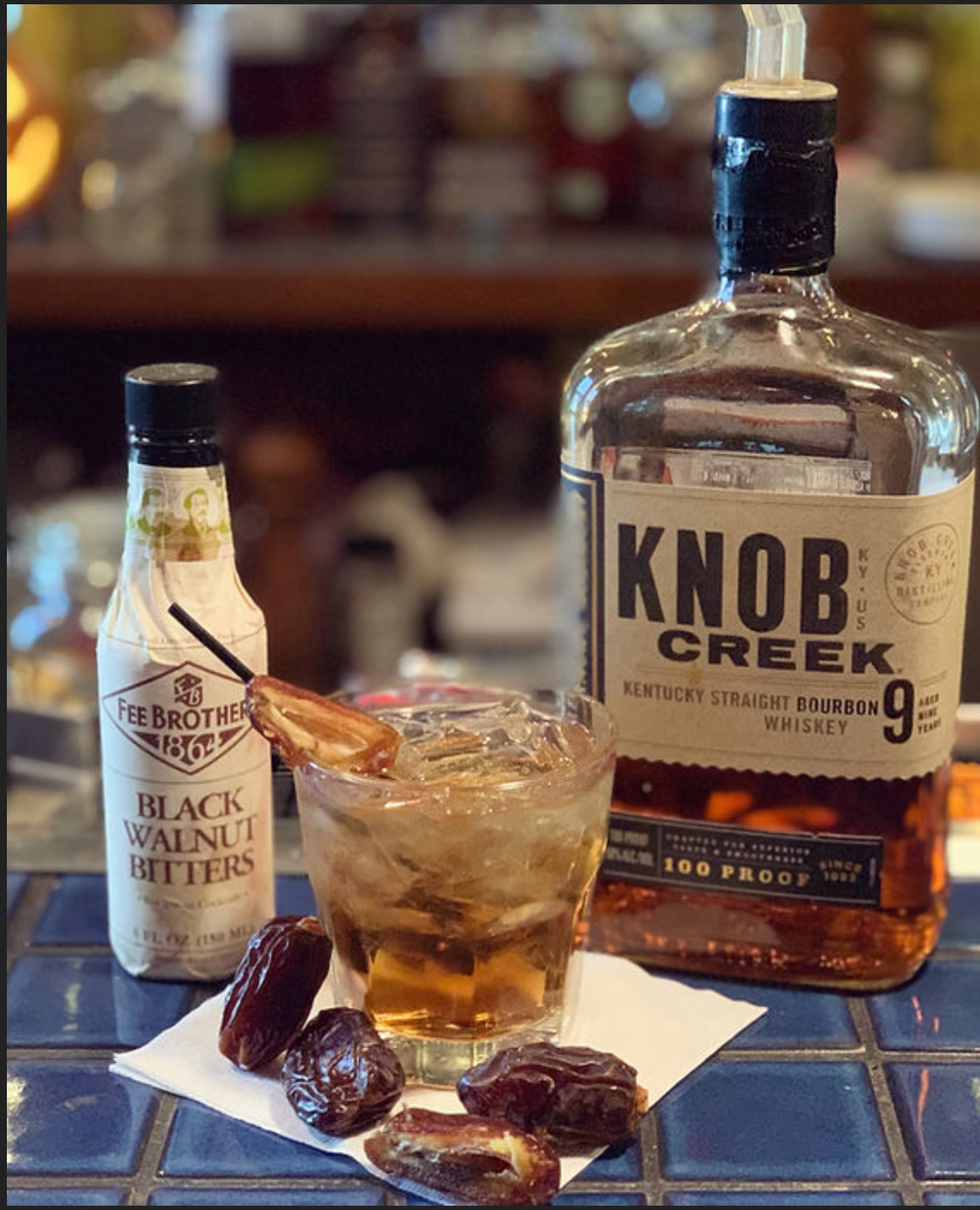 A bottle of knob creek whiskey next to a glass of whiskey and a bottle of black walnut bitters.