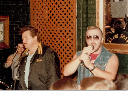 A man singing into a microphone wearing sunglasses and a jacket that says dan