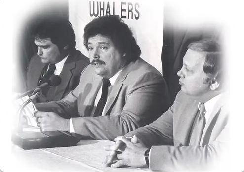 Three men in suits and ties are sitting at a table with microphones in front of a sign that says whalers