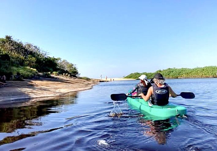 Duas pessoas remando em um rio calmo, um caiaque verde sob um céu azul claro.