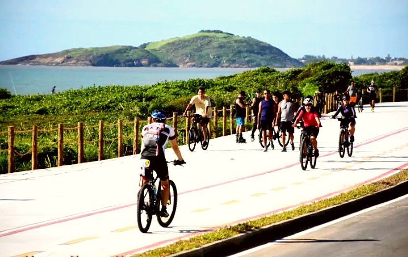Ciclistas percorrendo uma estrada costeira com o oceano e colinas verdes ao fundo.