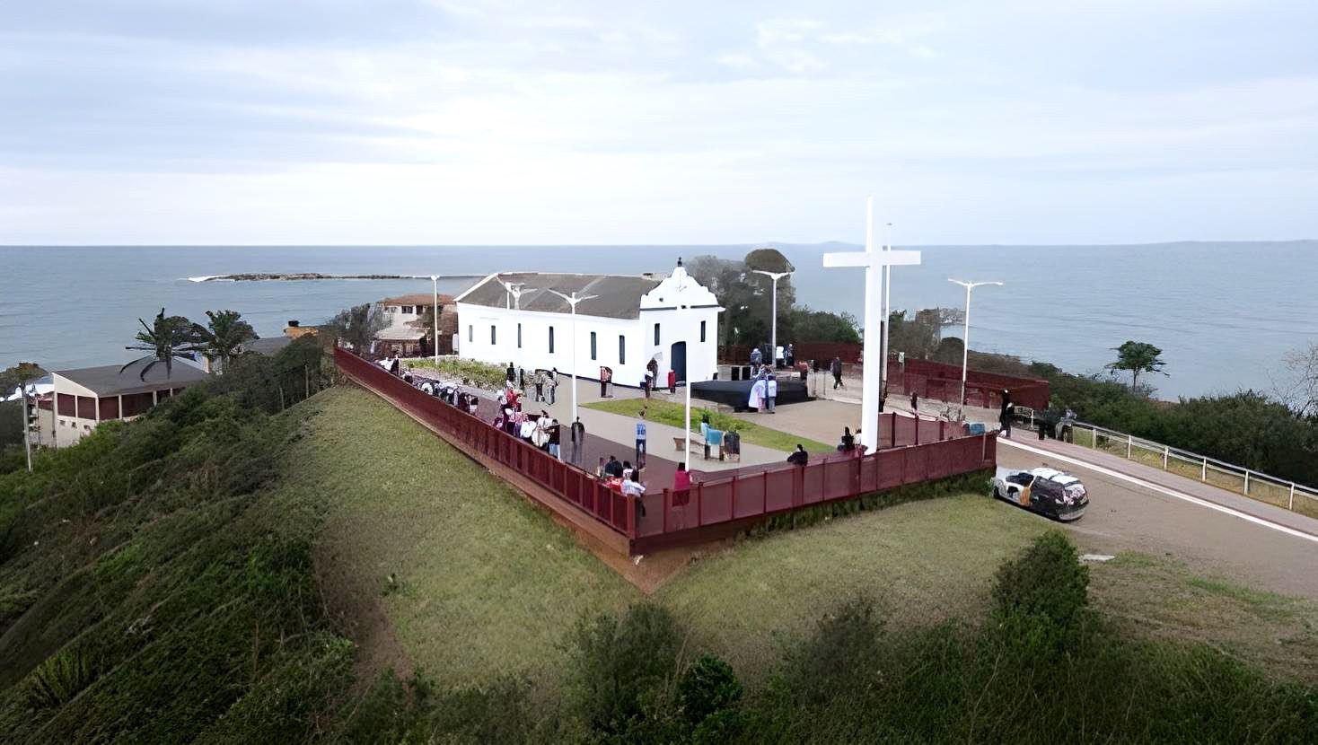 Igreja branca no alto de uma colina à beira-mar, com uma grande cruz e cerca vermelha com vista para o litoral.