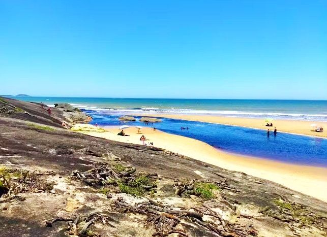 Costa rochosa com praia de areia, oceano azul e algumas pessoas sob um céu claro.