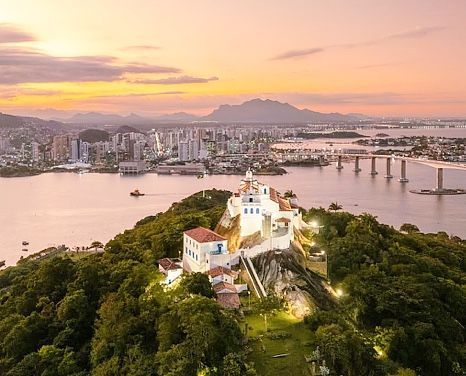 Pôr do sol sobre o porto da cidade, com uma igreja branca no topo de uma colina com vista para a água e o horizonte.
