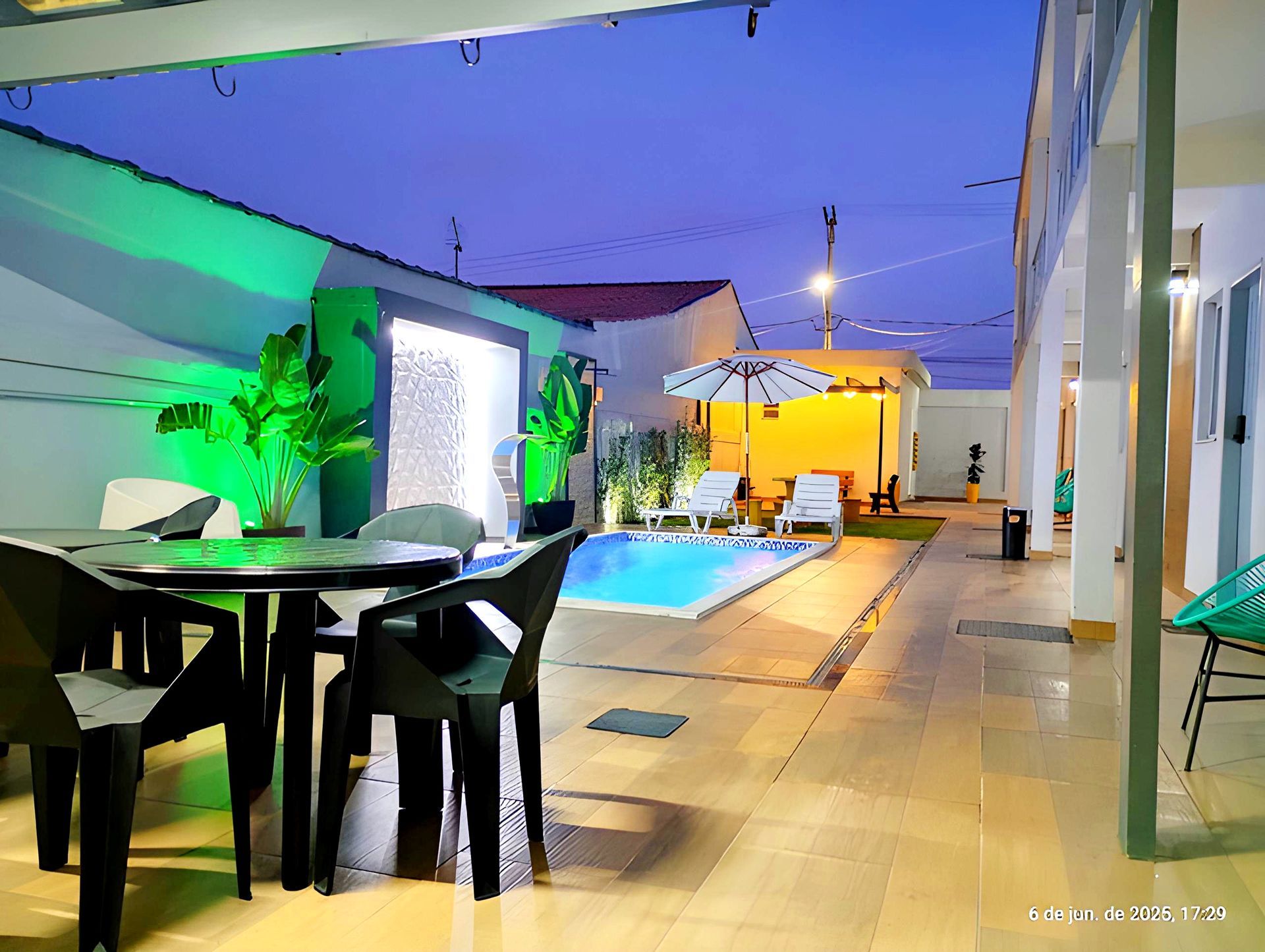 Pátio moderno com mesa de jantar, espreguiçadeiras, piscina e iluminação colorida ao entardecer.