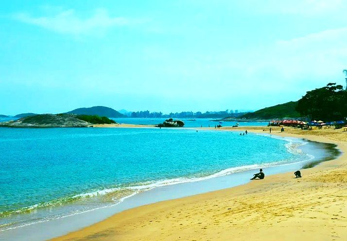 Praia tropical ensolarada com água turquesa, areia dourada e colinas verdes ao longe.