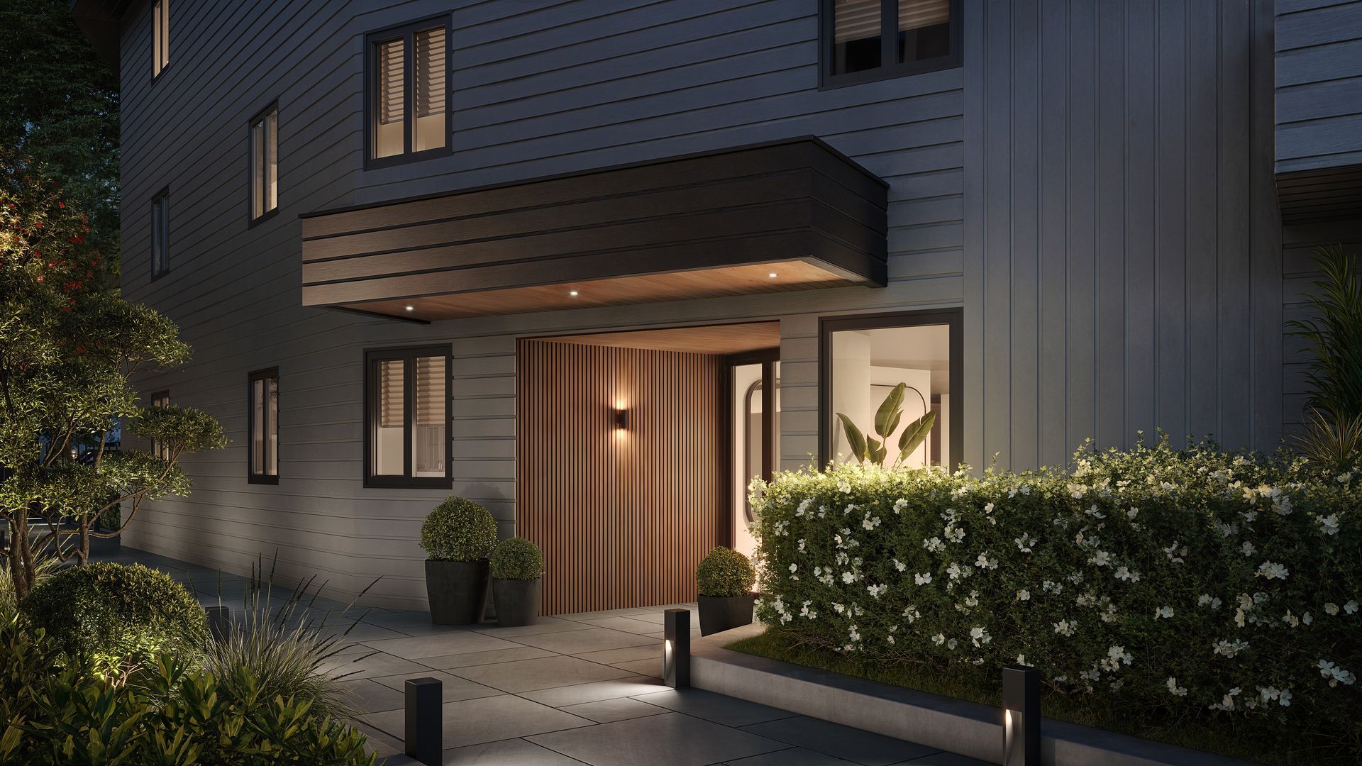 Nighttime exterior of a modern building with lit walkway, entrance, and lush landscaping.