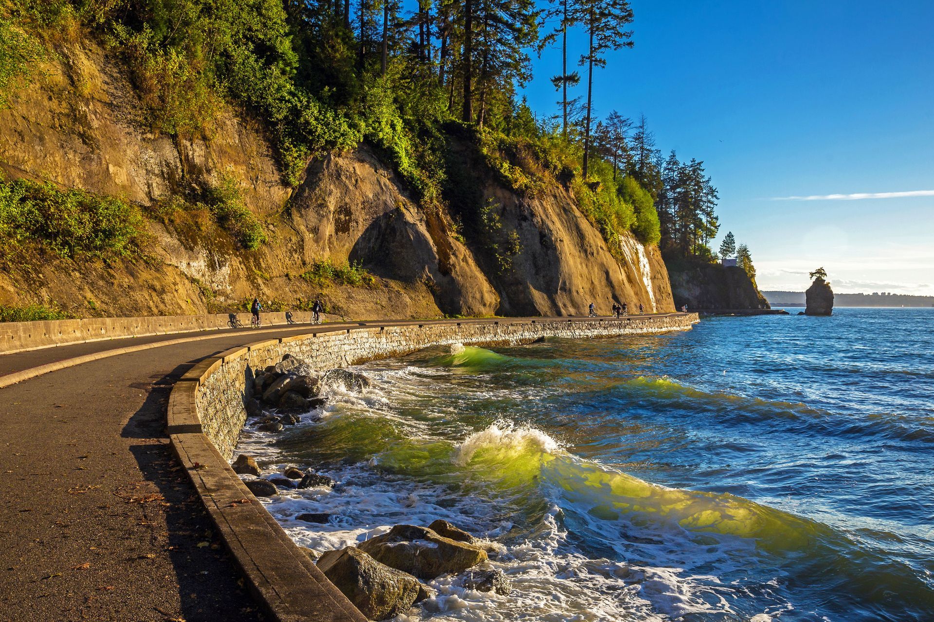 Paved path alongside a rocky coastline, water with waves, cliffside with trees, sunny sky.