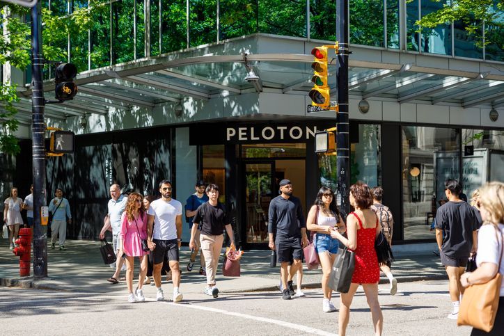 People crossing a street in front of a Peloton store, under a sunny sky.