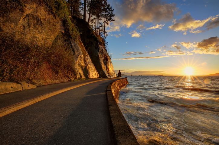 Paved road alongside ocean at sunset, illuminated by golden sunlight. Cliffs on left, sun on right.
