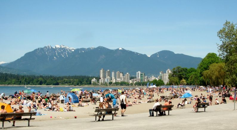 Beach scene with many people, mountains, and city skyline on a sunny day.