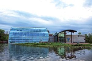 Building with glass walls on a lake, cloudy sky reflecting in the water.