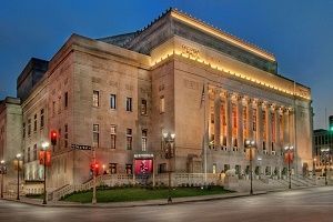Stone building with columns, lit at dusk. The building is on a city street corner.