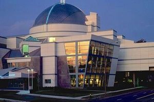 Modern building with a large blue dome and angled glass windows, lit up at dusk.
