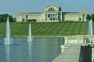 Museum building behind a lake with three fountains, on a sunny day.