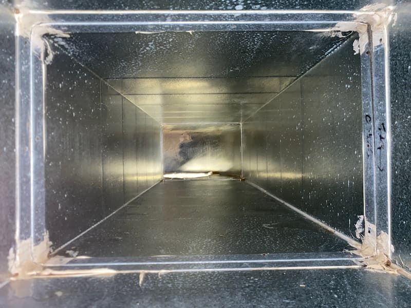 Inside view of a rectangular, metal air duct, appearing clean but with some debris visible.