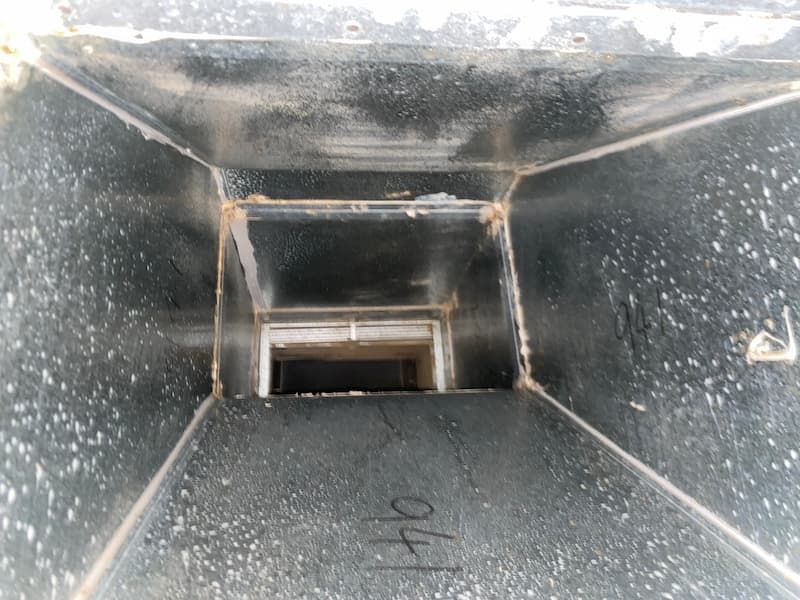 Inside view of a dusty metal air duct, appearing rectangular and converging towards a smaller opening.