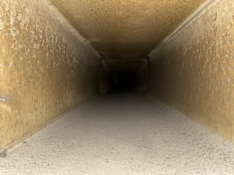 Interior of a rectangular air duct coated with a thick layer of dust and debris, with a dark, obscured center.