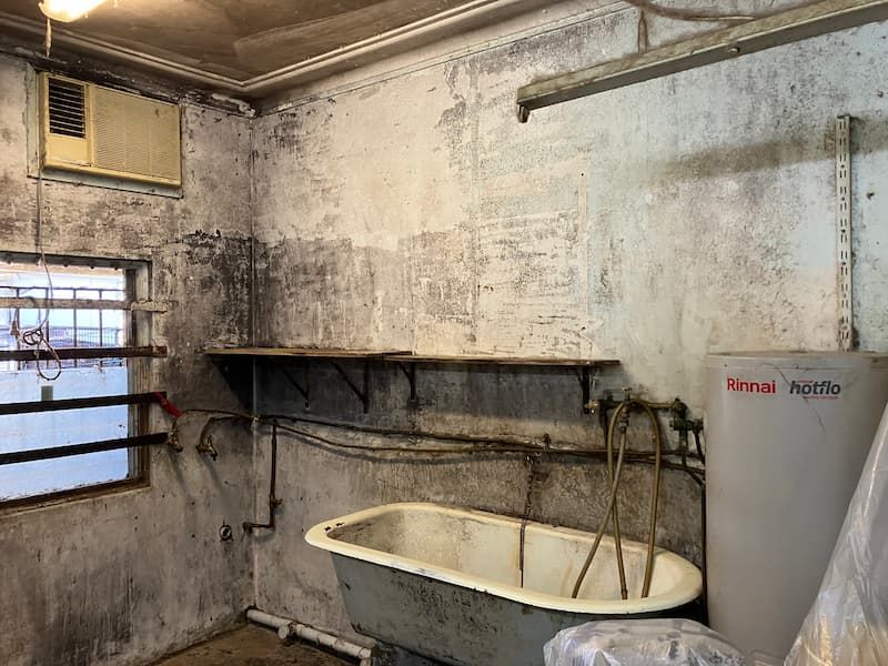 Moldy, dimly lit room with an old bathtub, shelving, and an air conditioner; a water heater is visible.