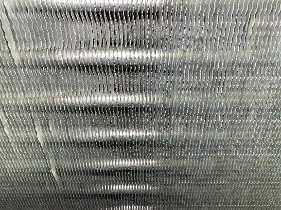 Close-up of a silver, metallic air conditioner coil with horizontal tubes and vertical fins.