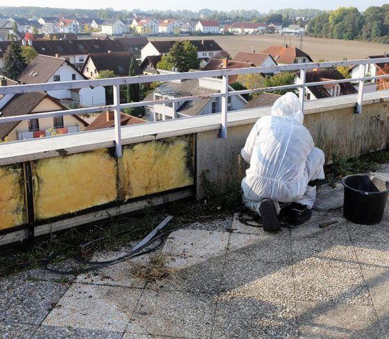 Person in protective suit kneeling on a rooftop, possibly removing something. Houses in the background.