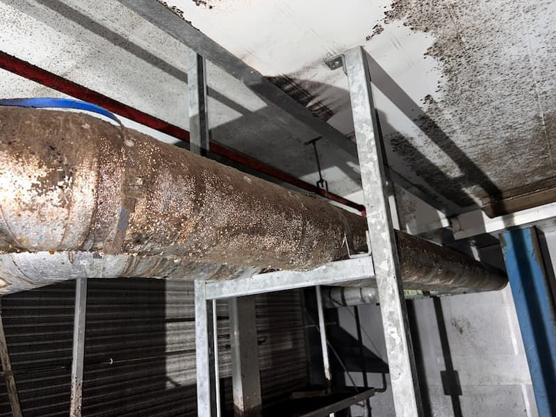 Corroded pipe in a room, supported by metal brackets. Ceiling shows water damage with brown and white stains.