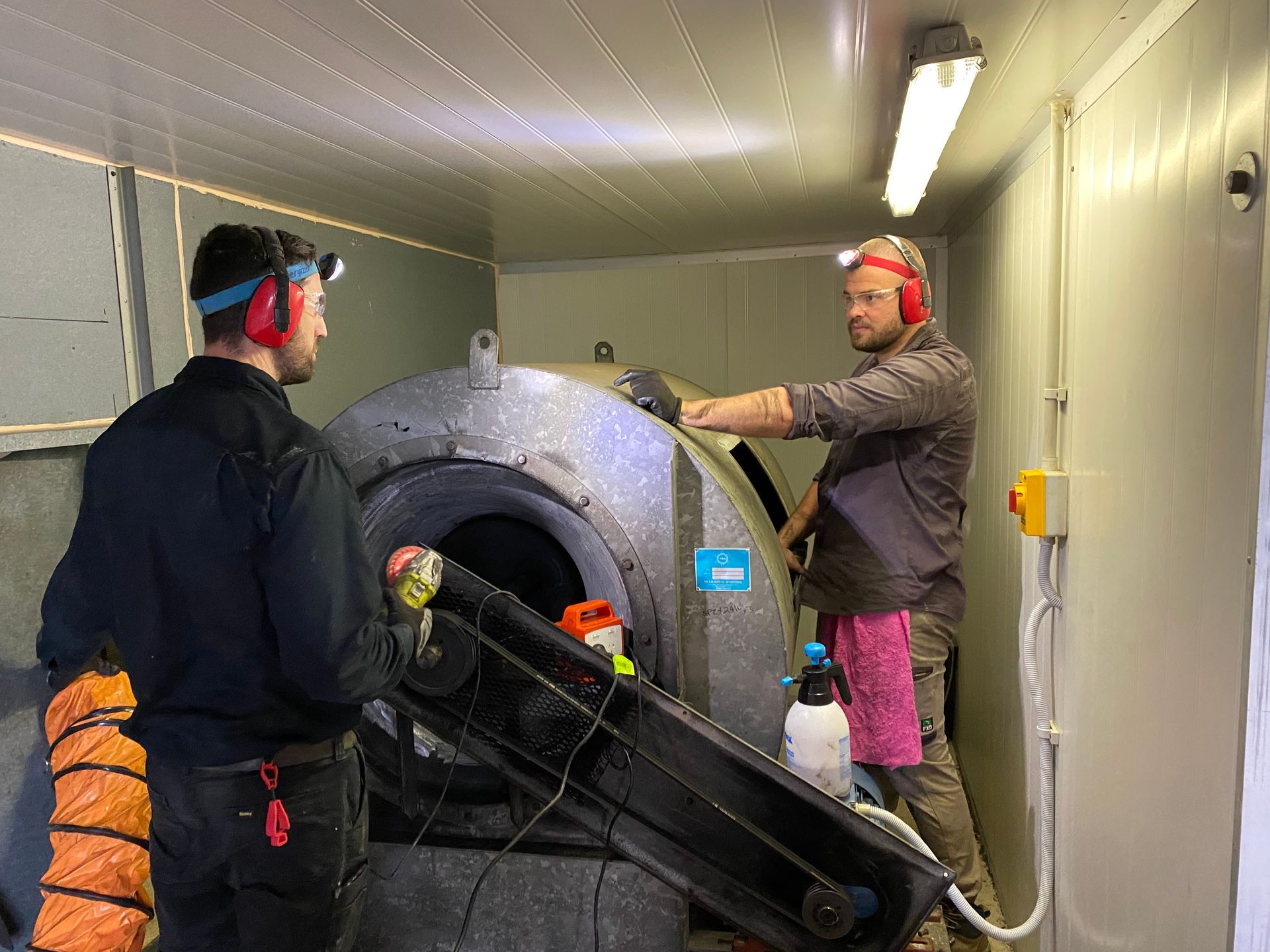 Two men working on machinery in an enclosed space; one holds a hose, both wearing ear protection and headlamps.