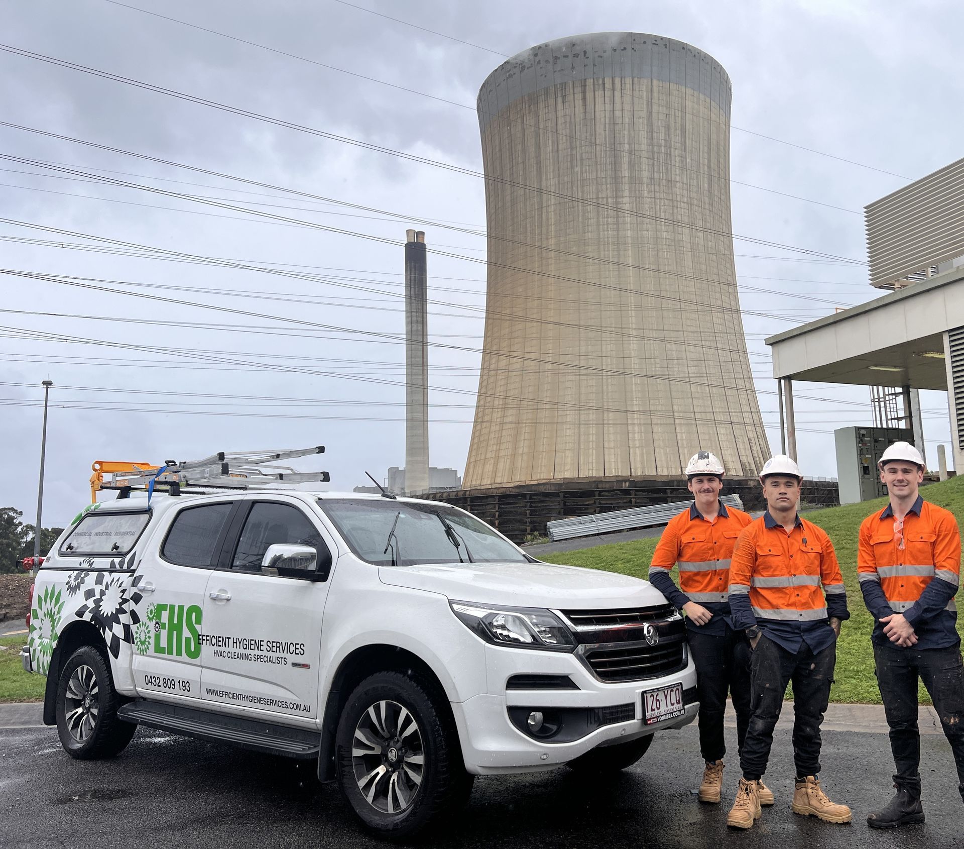 Three workers in orange work shirts and hard hats stand next to a white truck with a power plant in the background.