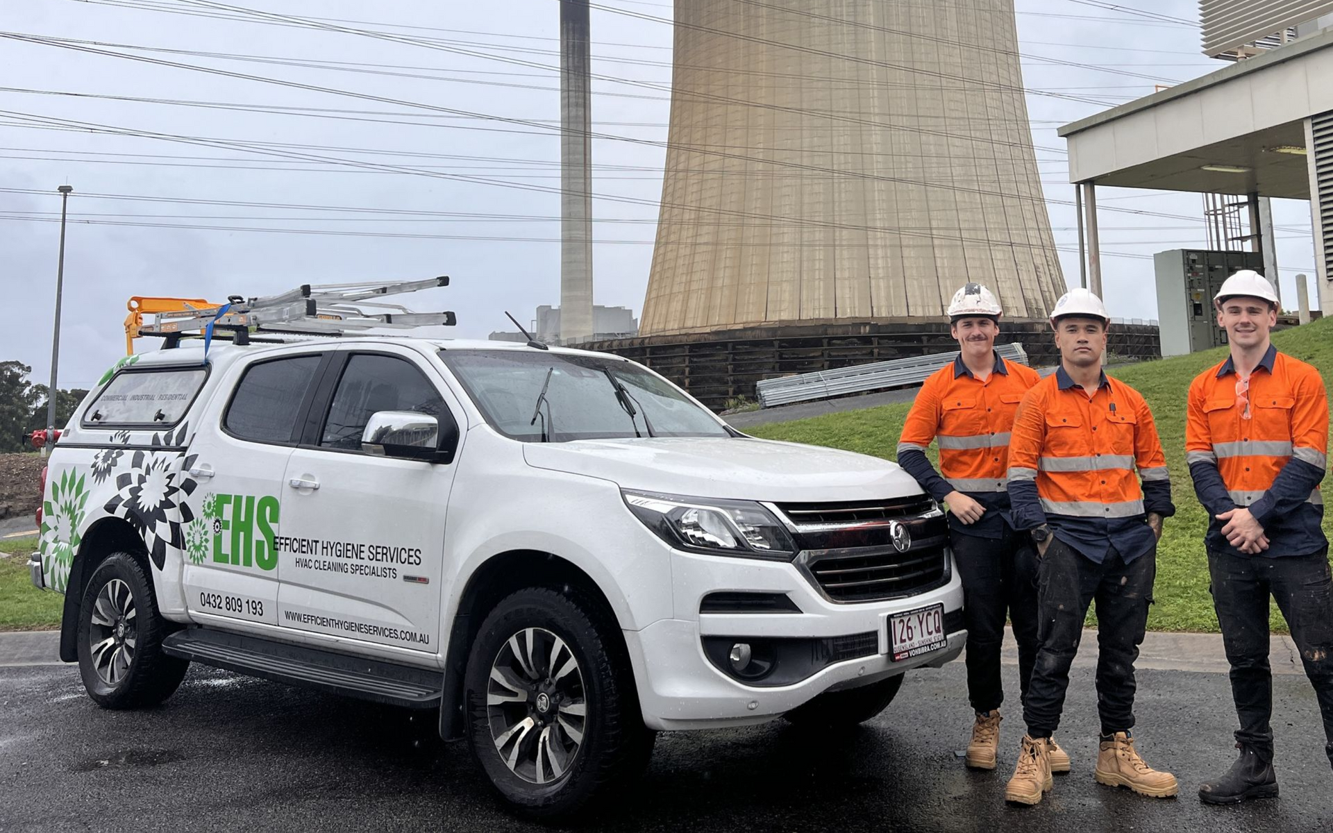 Three workers in safety gear pose beside a work truck with a power plant in the background.