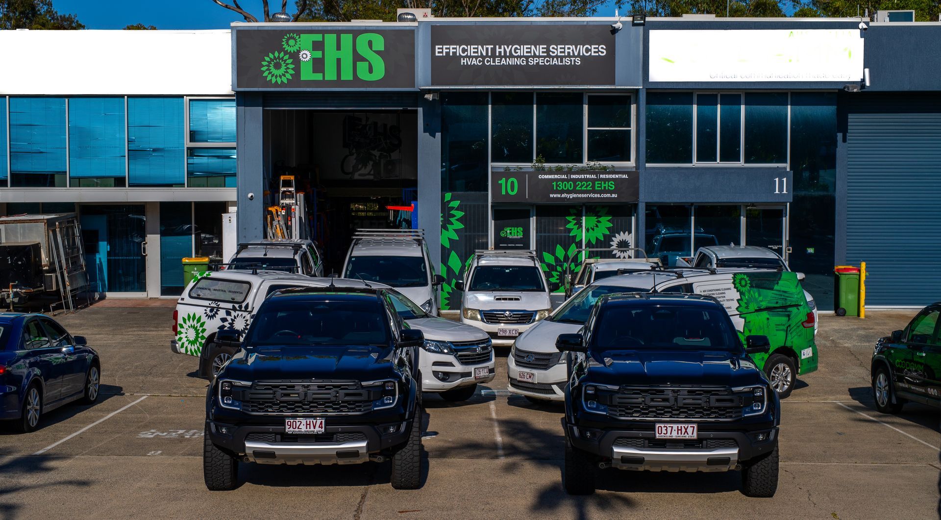 Two black trucks parked in front of a building with vans and cars. Building has signage and a green logo.