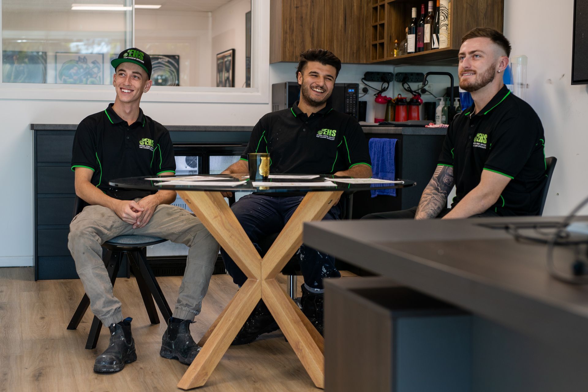 Three people sit around a table in an office, wearing black shirts with green accents, smiling.