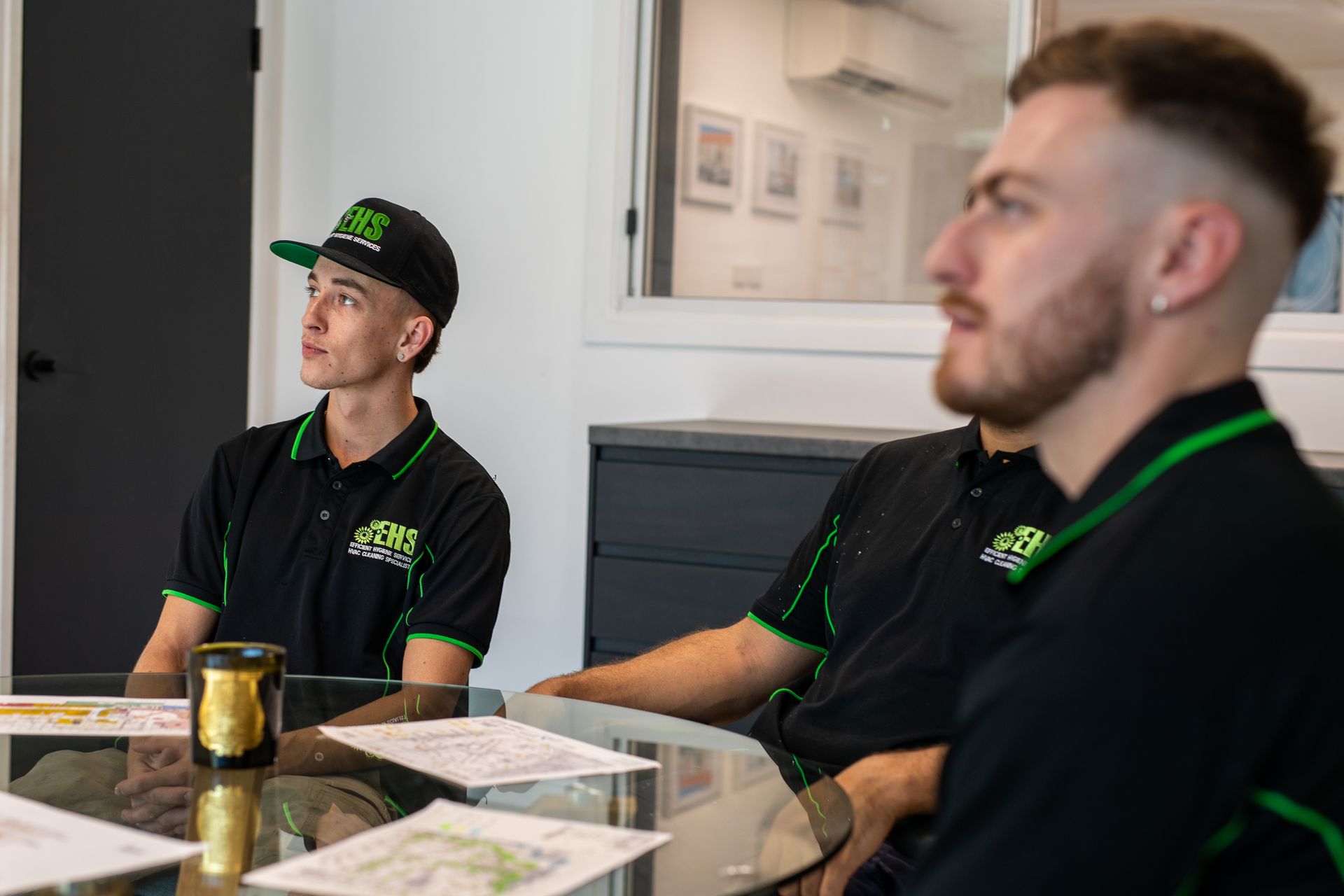 Three people in black shirts with green trim seated at a table, looking upwards in a bright office.