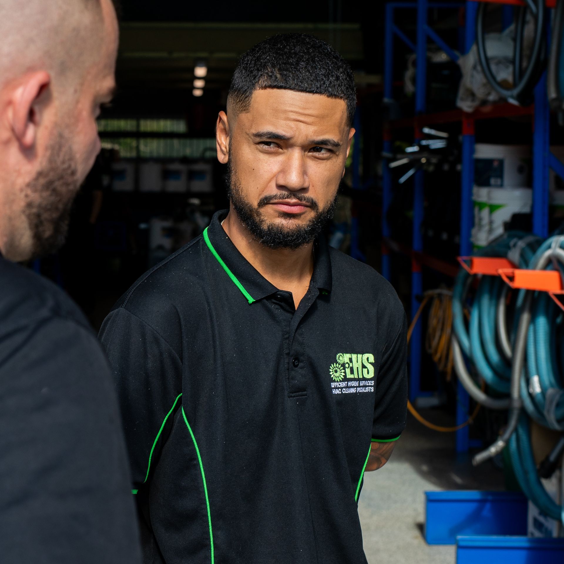 Man in black polo shirt, green trim, looking intently. Standing indoors near another man. Warehouse setting.