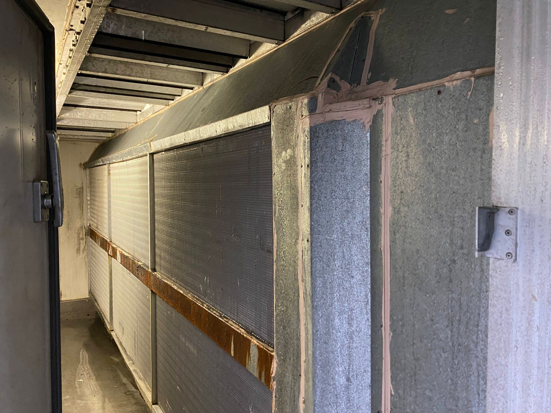Interior view of a long, narrow hallway. Gray metal and concrete construction with a grid pattern and brown support beams.