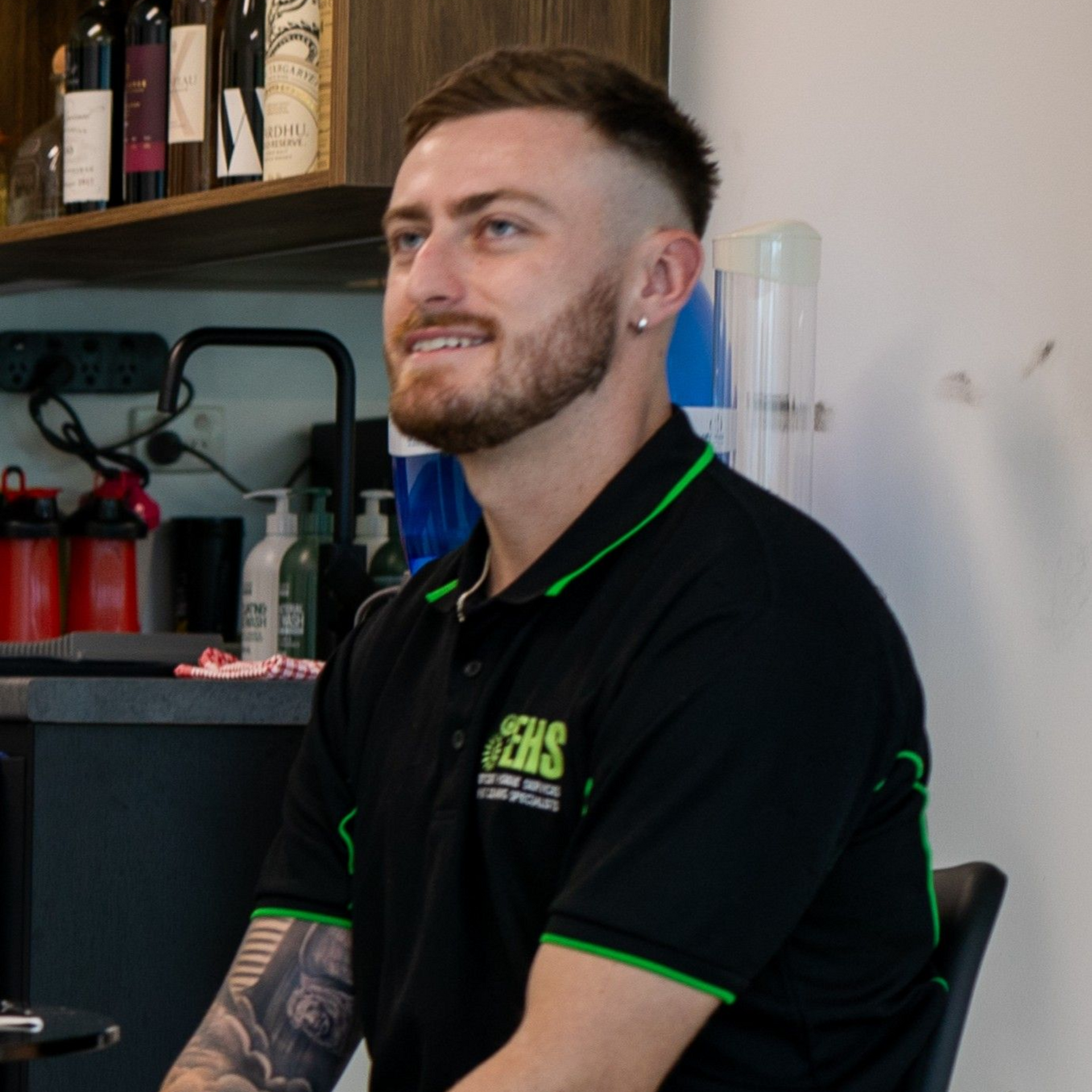 Man with short brown hair and beard, wearing a black shirt with green trim, smiling, inside.
