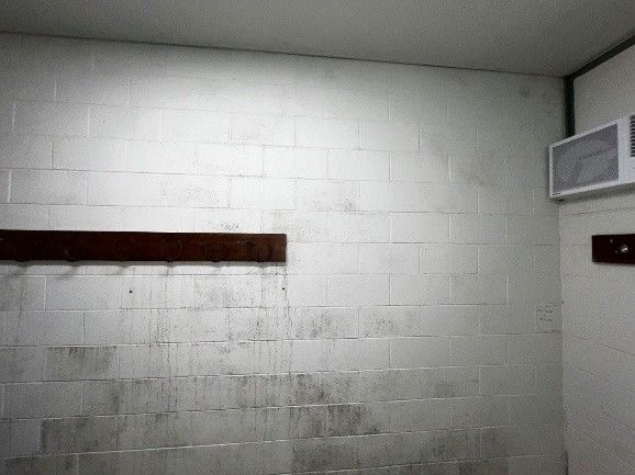 White brick wall with dark stains, a wooden shelf, and a ceiling-mounted unit.