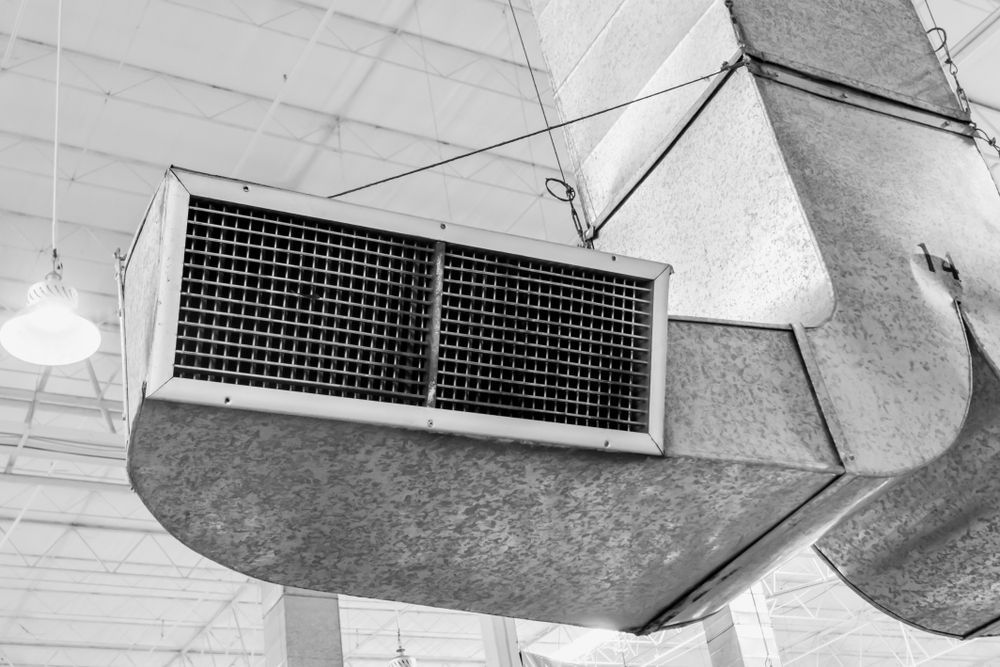 Grayscale view of industrial air duct with grilles, hanging from a warehouse ceiling.