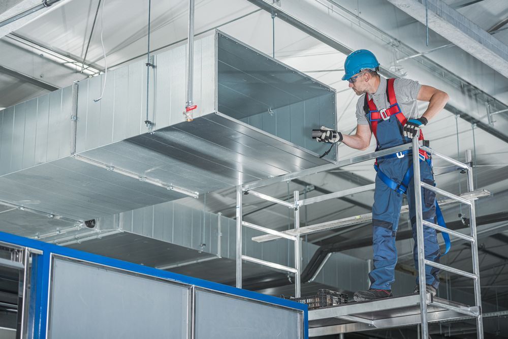 HVAC technician inspects ductwork from scaffolding, wearing a hard hat and safety harness in a commercial building.