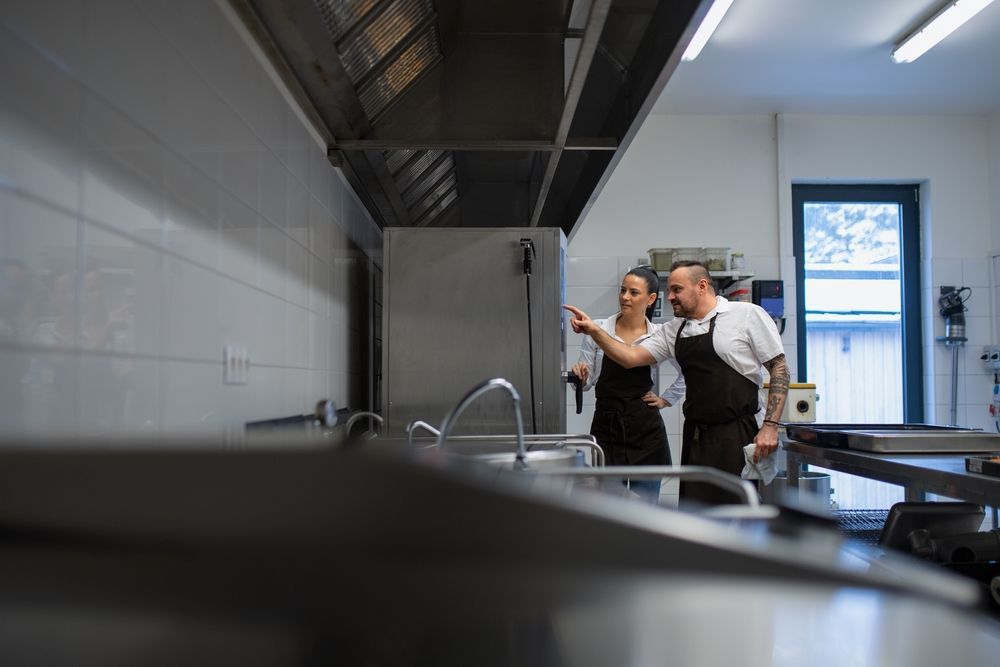 Two chefs in aprons discussing in a commercial kitchen; one points upward near a ventilation hood.