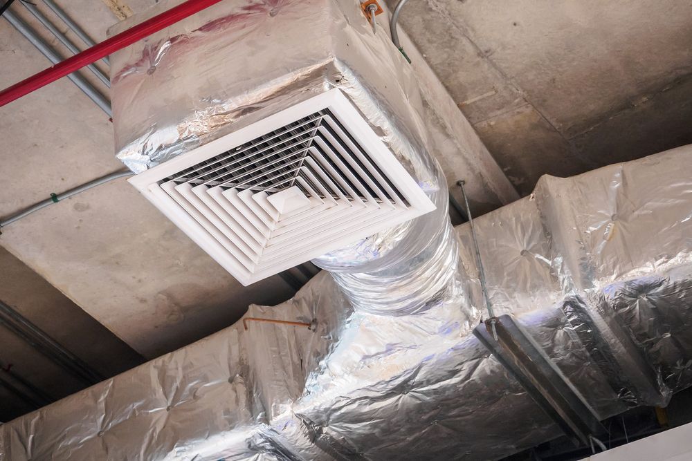 Air vent on a ceiling. The square vent is white, surrounded by ductwork wrapped in foil insulation.