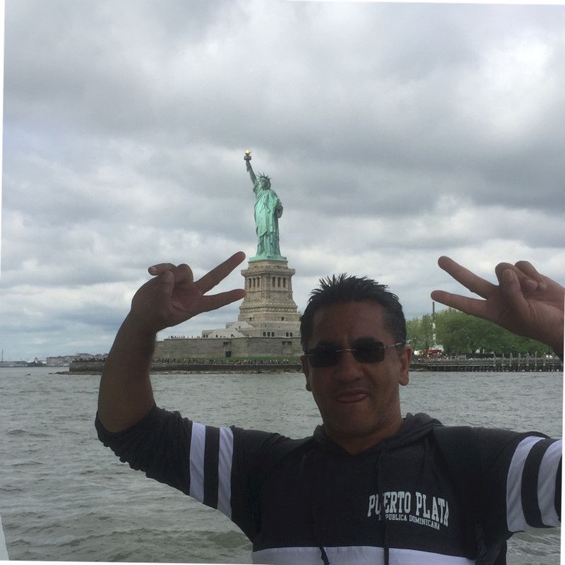 Man makes peace signs in front of the Statue of Liberty. Cloudy sky, water.