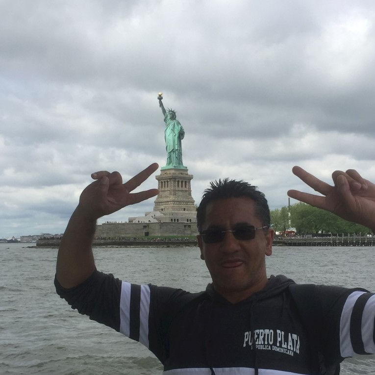Man makes peace signs in front of the Statue of Liberty. Cloudy sky, water.