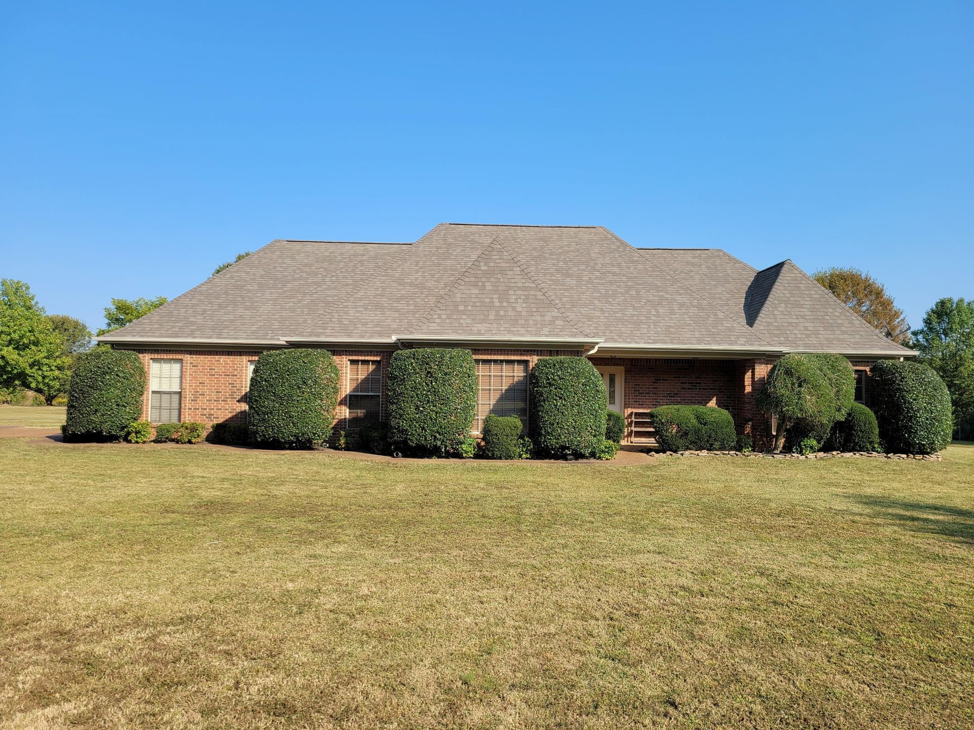 Red brick ranch-style house with brown roof, surrounded by green bushes and a large grassy yard under a blue sky.