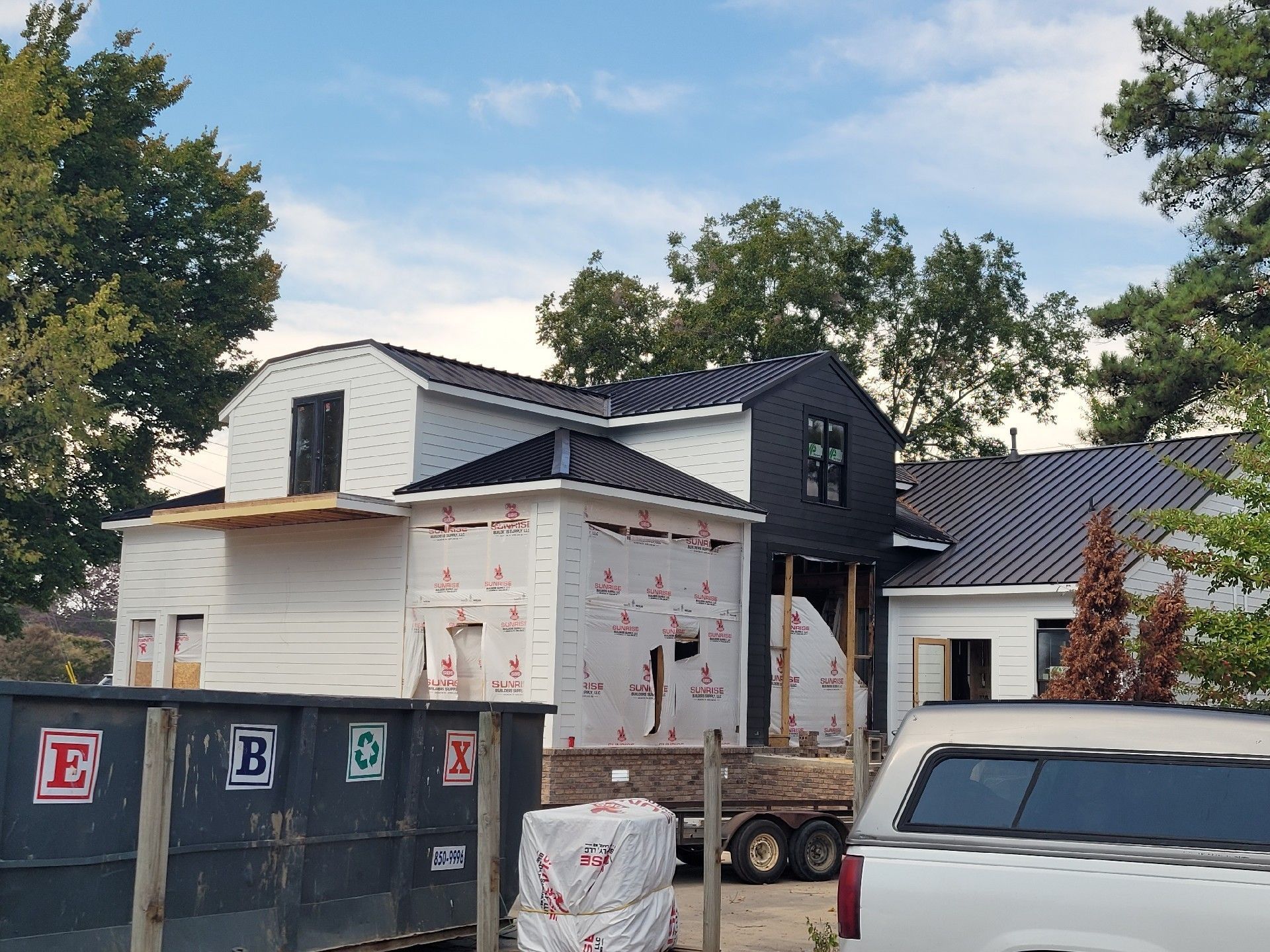 House under construction with black roof, white siding, surrounded by trees, and a dumpster in front.