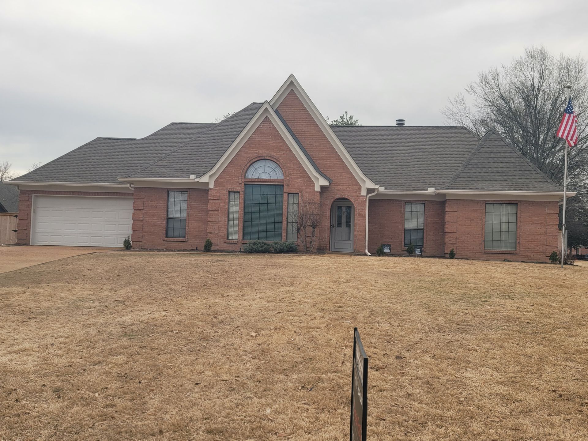 Red brick house with brown roof, garage, and American flag on a dry lawn.